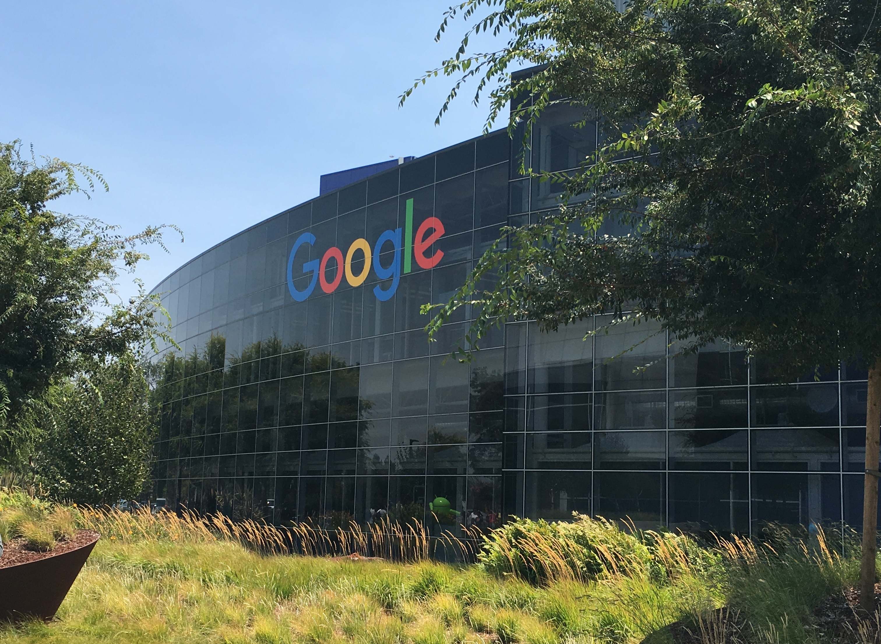Logo Google sur la façade vitrée de son siège à Mountain View, entourée d'arbres.