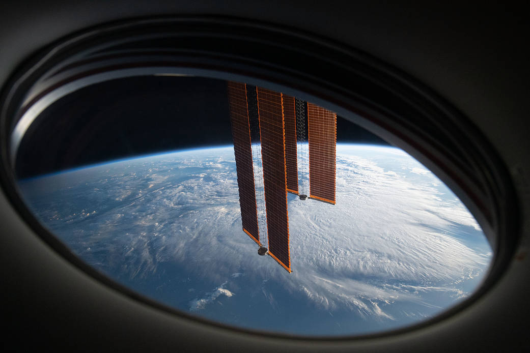 Deux panneaux solaires de la Station spatiale internationale devant la Terre, vus à travers un hublot.